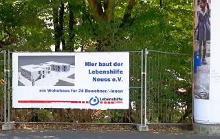 Baustellenplakat mit der Aufschrift: " Hier baut der Lebenshilfe Neuss E.V. ein Wohnhaus für 24 Bewohner/-innen.