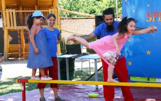 Kinder führen eine Zirkus-Nummer auf, auf dem Sommerfest im Familienzentrum Am Baldhof. Zwei Mädchen balancieren mit Hilfestellung durch einen Erwachsenen auf einem Balken.