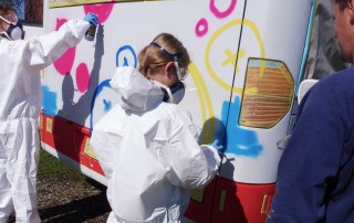 Zwei Personen in weißen Schutzanzügen besprühen den Jugendbus mit Graffiti-Farbe um ihn neu zu gestalten.