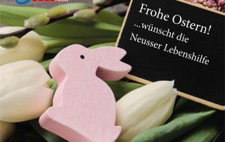 Ein rosa Hase aus Holz liegt zwischen Tulpen. Daneben ist eine Tafel auf der steht: "Frohe Ostern! ...wünscht die Neusser Lebenshilfe"