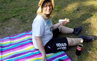 Eine junge Frau sitzt auf einer Picknick-Decke auf einer Wiese. Sie isst einen Hot-Dog, trägt einen Sonnenhut und lächelt in die Kamera.