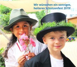 Porträt eines Mädchen und eines Jungen die beide Hüte tragen, das Mädchen hält eine Blume in der Hand. Auf dem Bild steht der Text: Wir wünschen ein sonniges, heiteres Schützenfest 2019!