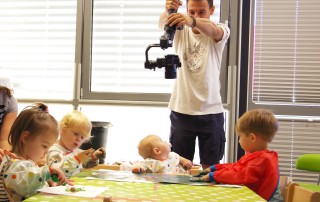 Ein Mann filmt mit einer Kamera eine Gruppe Kindergartenkinder die beim Essen an einem Tisch sitzen.