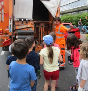 Ein Mitarbeiter der Müllentsorgung steht hinter einem Müllauto und erklärt einer Gruppe Kinder etwas.