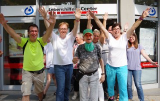 Eine Gruppe Mitarbeitende der Lebenshilfe Neuss und Nutzer stehen vor dem Lebenshilfe Center und reißen fröhlich die Arme nach oben.