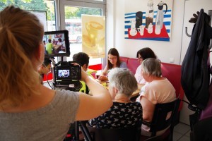 Eine Frau mit einer Fernsehkamera filmt eine Gruppe Nutzerinnen des Lebenshilfe Centers, die an einem Tisch sitzen.