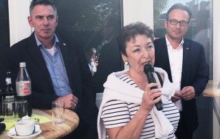 Angelika Quiring-Perl spricht in ein Mikrofon. Hinter ihr stehen zwei Männer die zuhören.