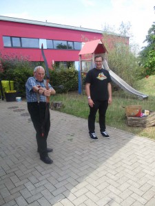 Ein alter Mann schießt bei einem Sommerfest der UWOs der Lebenshilfe Neuss mit Pfeil und Bogen. Ein jüngerer Mann steht daneben und sieht ihm zu.