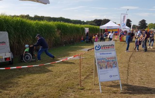Das Familienfest im Dycker Feld. Im Hintergrund sieht man Menschen die das Fest besuchen und den Stand der Lebenshilfe Neuss. Im Vordergrund steht eine Werbetafel die auf den Stand der Lebenshilfe aufmerksam macht.