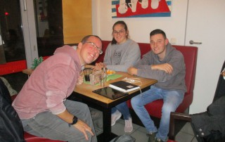 Drei Nutzer und Mitarbeiter der Lebenshilfe Neuss sitzen im Lebenshilfe Center beim Tisch-Kicker Turnier an einem Tisch und lachen in die Kamera.