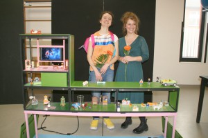 Zwei Frauen mit jeweils einer Rose in der Hand stehen hinter einem Tisch auf dem Playmobilfiguren sind.