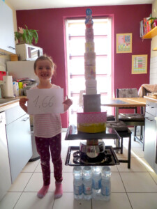 Ein Mädchen steht in einer Küche neben einem selbst gebauten Turm. Sie hält einen Zettel in der Hand auf dem 1,66 (Meter) steht.