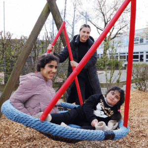 Zwei Kinder sitzen in einer runden Schaukel und werden von einer Frau angeschoben.
