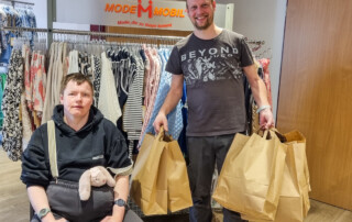 Zwei Personen stehen vor einer Kleider-Stange. Eine Person trägt Tüten