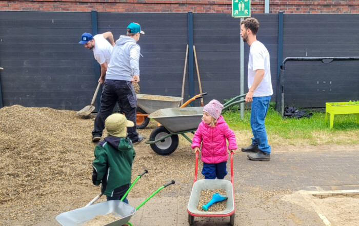 Zwei Kinder und drei Väter schaufeln Kies in eine Schubkarre und schieben diese weg.