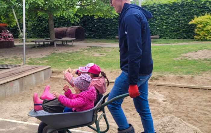 Ein Mann schiebt zwei Kinder in einer Schubkarre vor sich her.