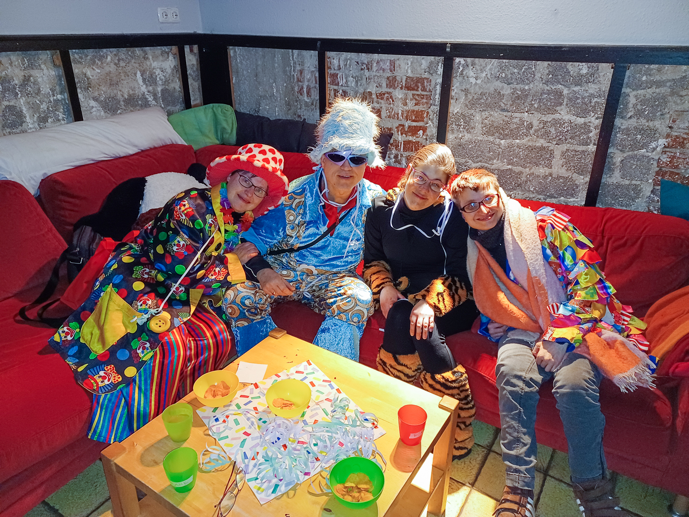 2505–15-UK-Karnevalsparty-OH‑1 Vier Besucher einer Party der offenen Hilfen der Lebenshilfe Neuss. Sie tragen bunte Karnevals-Verkleidungen und sitzen auf einer roten Eck-Couch und lächeln in die Kamera.