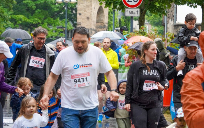 Eine große Gruppe Menschen läuft beim Sommernachtslauf in Neuss 2025. Im Fokus ein Mann in einem weißen Oberteil mit dem Logo der Lebenshilfe Neuss. An seiner Hand hält er ein Kind, das ebenfalls läuft und auch ein Oberteil der Lebenshilfe Neuss trägt.