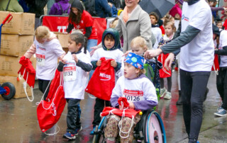 Eine Gruppe Kinder in weißen T-Shirts mit dem Logo der Lebenshilfe Neuss läuft beim Sommernachtslauf. Eines der Kinder sitzt im Rollstuhl und wird von einem Mann geschoben.