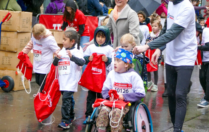 Eine Gruppe Kinder in weißen T-Shirts mit dem Logo der Lebenshilfe Neuss läuft beim Sommernachtslauf. Eines der Kinder sitzt im Rollstuhl und wird von einem Mann geschoben.