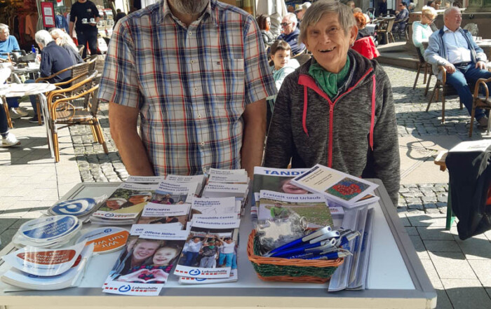 Eine Frau und ein Mann stehen hinter einem Stand der Lebenshilfe Neuss auf einem Stadt-Fest und verteilen Infomartions-Material.