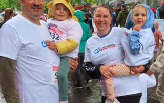 Gruppenfoto einer Mutter und eines Vaters, die beide ein Mädchen auf dem Arm tragen. Sie haben alle weiße T-Shirts der Lebenshilfe Neuss an und lachen in die Kamera.