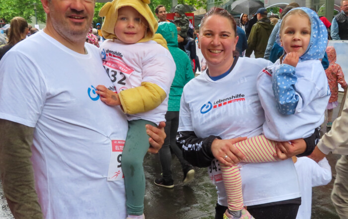 Gruppenfoto einer Mutter und eines Vaters, die beide ein Mädchen auf dem Arm tragen. Sie haben alle weiße T-Shirts der Lebenshilfe Neuss an und lachen in die Kamera.