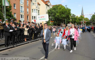Die Kinder laufen in ihren Kostümen den Umzug entlang. Am Anfang hält ein Mann ein Schild des Familienzentrum Marienburg hoch.