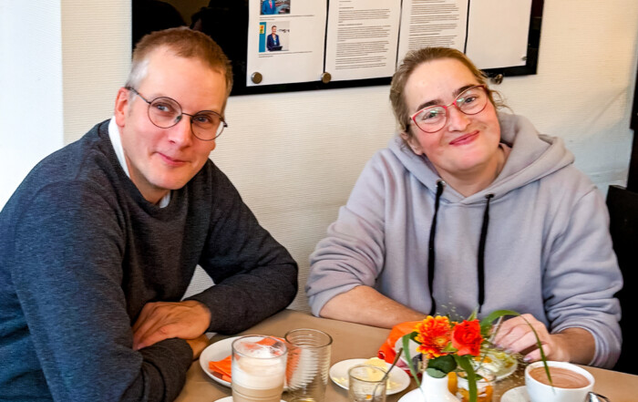 Zwei Personen sitzen am Tisch und lächeln in die Kamera.