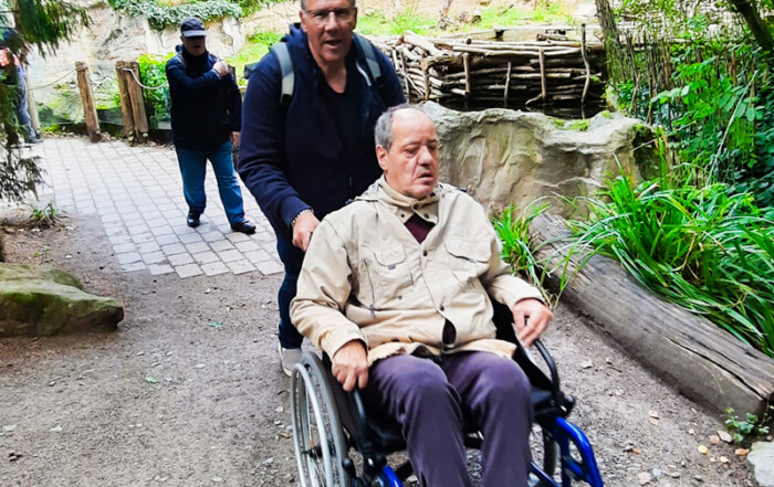 Ein Mann im Rollstuhl wird von einem Betreuer durch den Zoo begleitet.