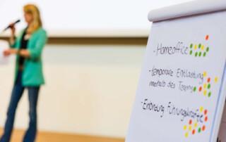 Aufnahme einer Rednerin und im Vordergrund stehen Notizen an einem Whiteboard.