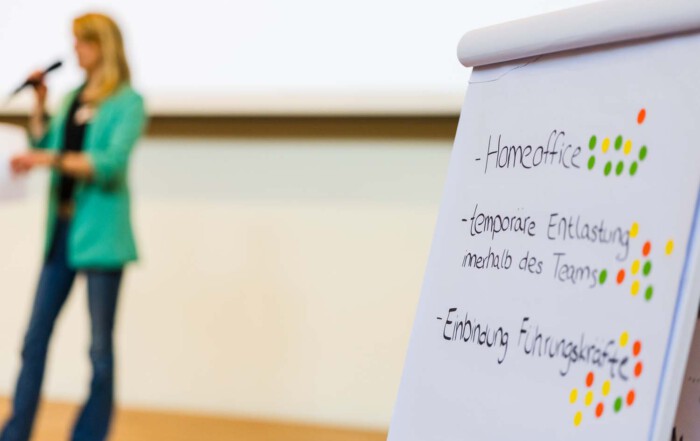 Aufnahme einer Rednerin und im Vordergrund stehen Notizen an einem Whiteboard.